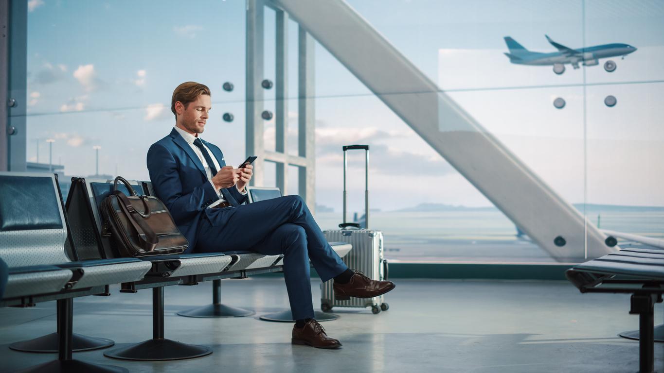 Airport Terminal: Businessman Uses Smartphone, Waiting for a Flight, Doing e-Business, Sending e-Commerce Data. Traveling Man Remote Work Online on Mobile Phone in Boarding Lounge of Airline Hub